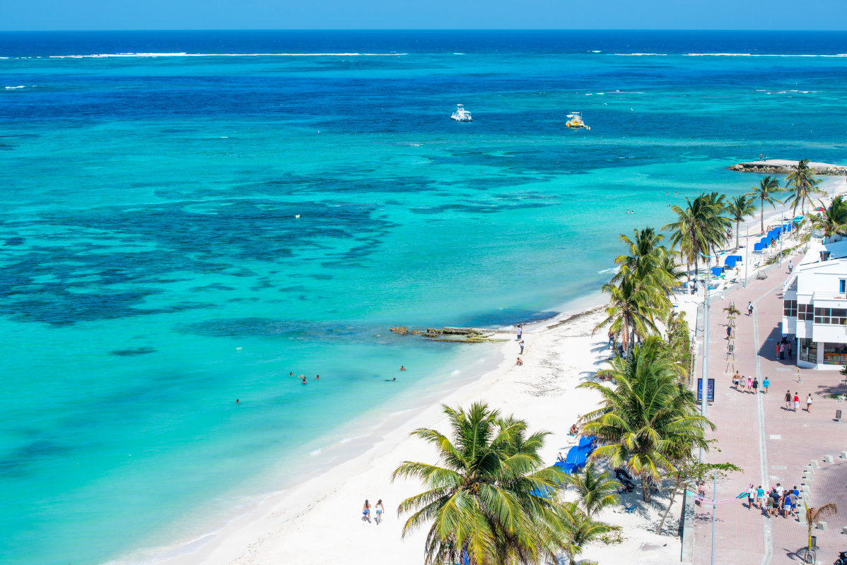 Aerial view of San Andres Island's shoreline featuring a bustling beachfront promenade lined with palm trees, beachgoers, and clear azure waters, which are just some things you can expect from this guide to San Andres, Colombia. 