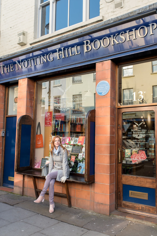 A young woman sitting outside the iconic Notting Hill Bookshop in London, a charming spot for book lovers and a quintessential stop for those visiting the famous Notting Hill area, adding to the literary charm of London.