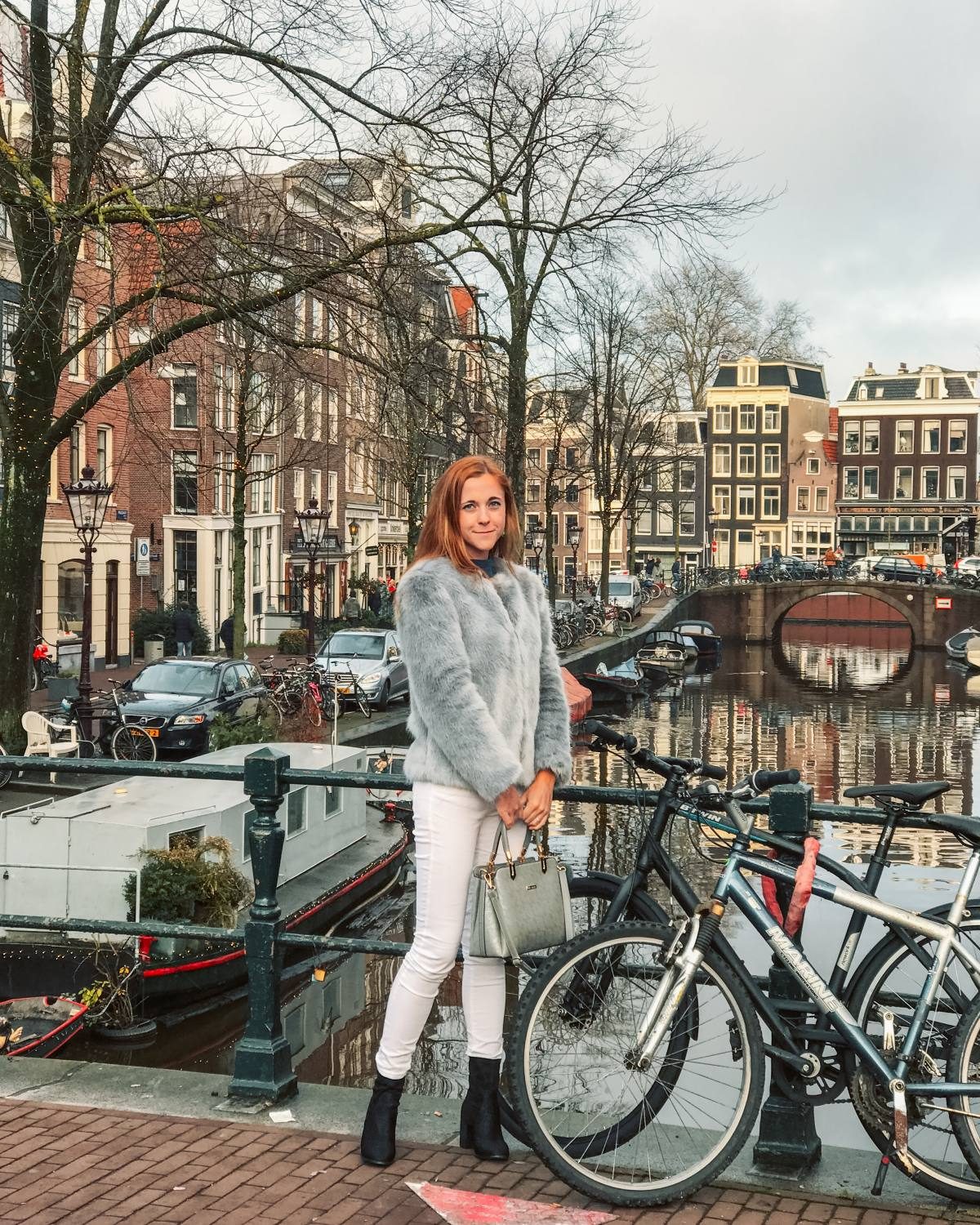 Woman posing in front of a picturesque Amsterdam canal with bikes and historical architecture, showcasing the charm of staying in the city's core.
