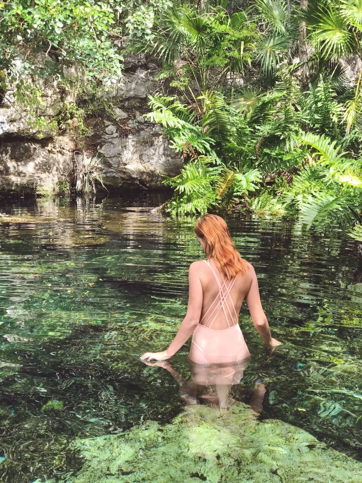 Lady wearing a nude-colored one piece suit entering a river.