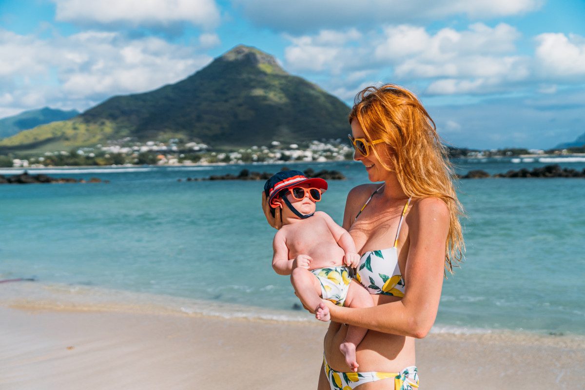 A woman holds small baby at a beach on their first holiday with a baby