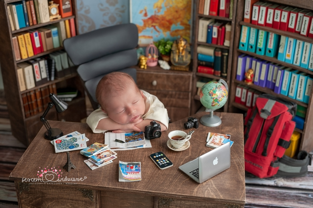 Baby dressed as a businessman at a desk covered with travel items and a laptop, humorously suggesting starting Spanish young.
