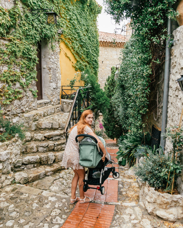 Mother carrying a stroller down an enchanting cobblestone alley overgrown with greenery, epitomizing the charming experiences of France with a baby.
