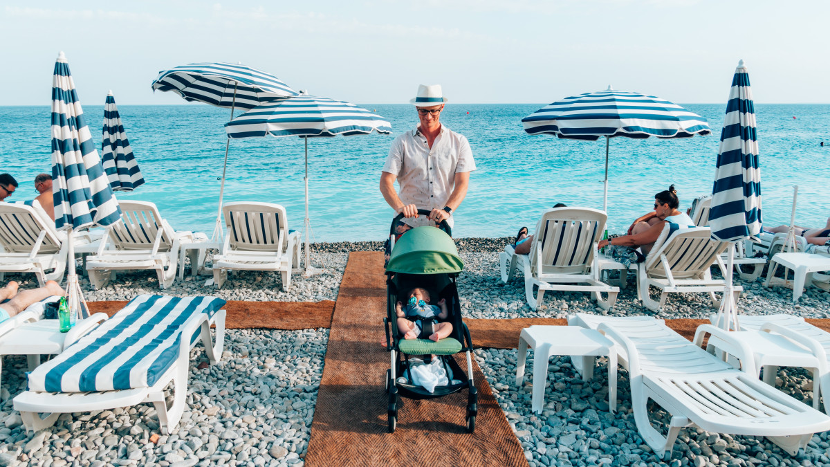 Father pushing the Babyzen Yoyo+ travel stroller along a pebble beach with beach chairs and umbrellas, epitomizing a family-friendly vacation accessory.
