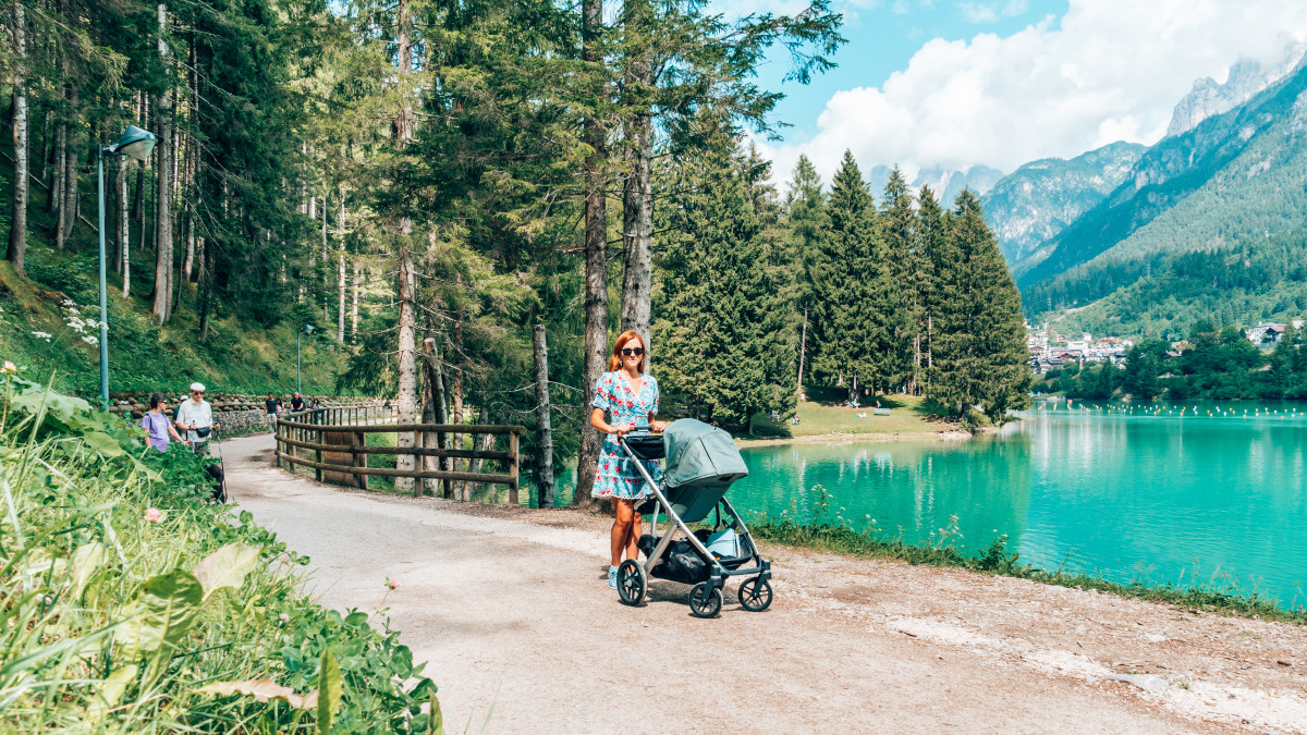 Woman strolling with a baby in a stroller along a picturesque lakeside path surrounded by mountains, perfect for travel.