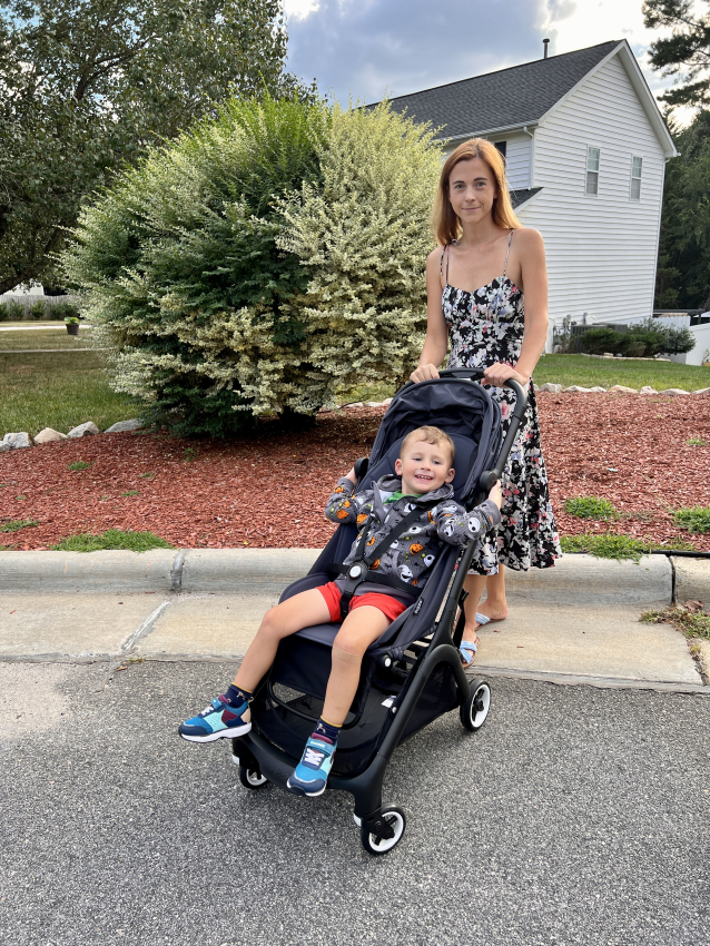 Happy toddler in a black stroller with a playful monster print hoodie, accompanied by a woman in a floral dress.
