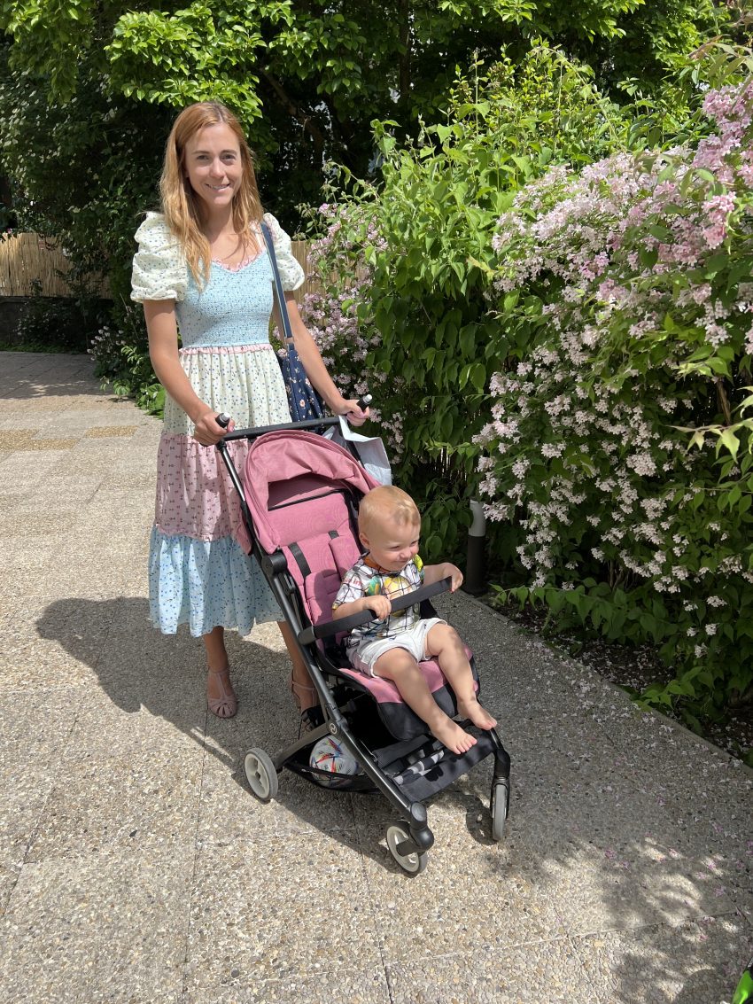 Mother strolling with a pink compact travel stroller, suitable for airplane overhead storage, through a blooming garden.