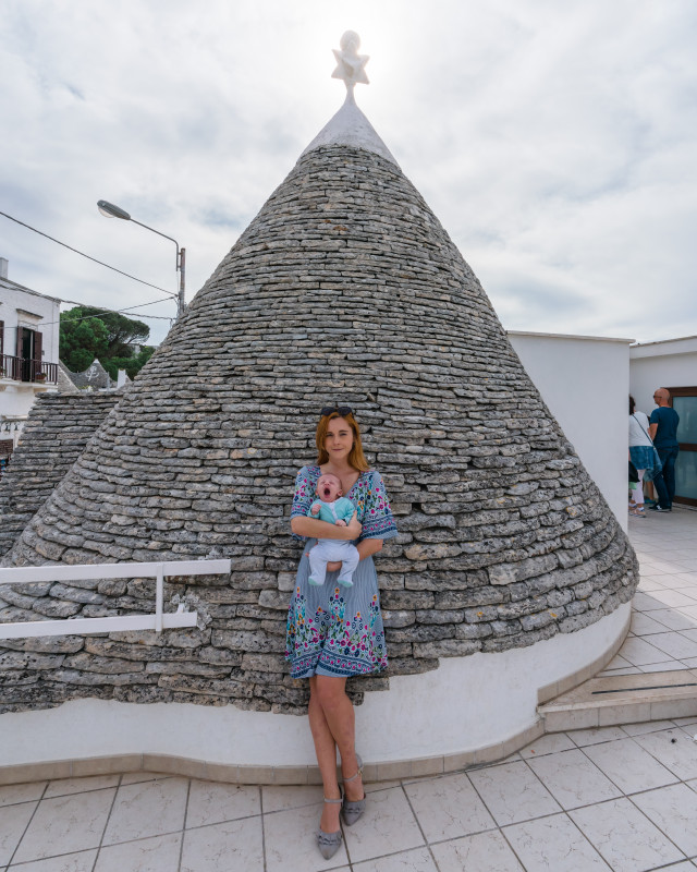 Woman cradling a baby against the unique backdrop of Alberobello’s trulli houses, exemplifying Italy’s diverse travel experiences with a child.
