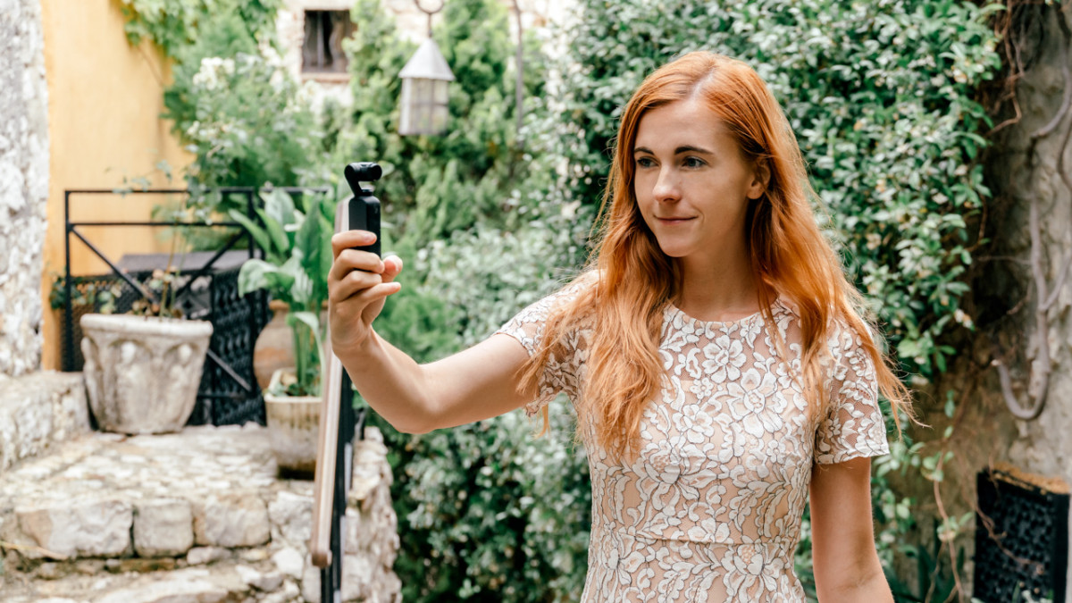 Red-haired woman using a video recorder while exploring a quaint garden setting.




