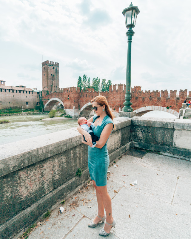 Young mom cradles her baby in front of the medieval Castelvecchio Bridge in Verona, highlighting the ease of exploring historic sites with a baby.