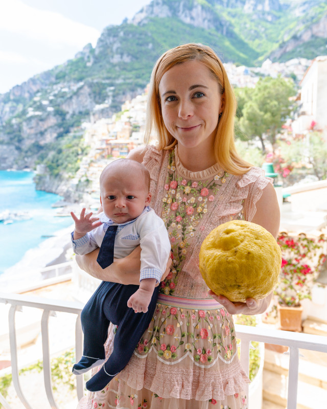 Smiling woman holding a baby and a lemon on a balcony overlooking the Amalfi Coast, embodying the joys of traveling to Italy with an infant.
