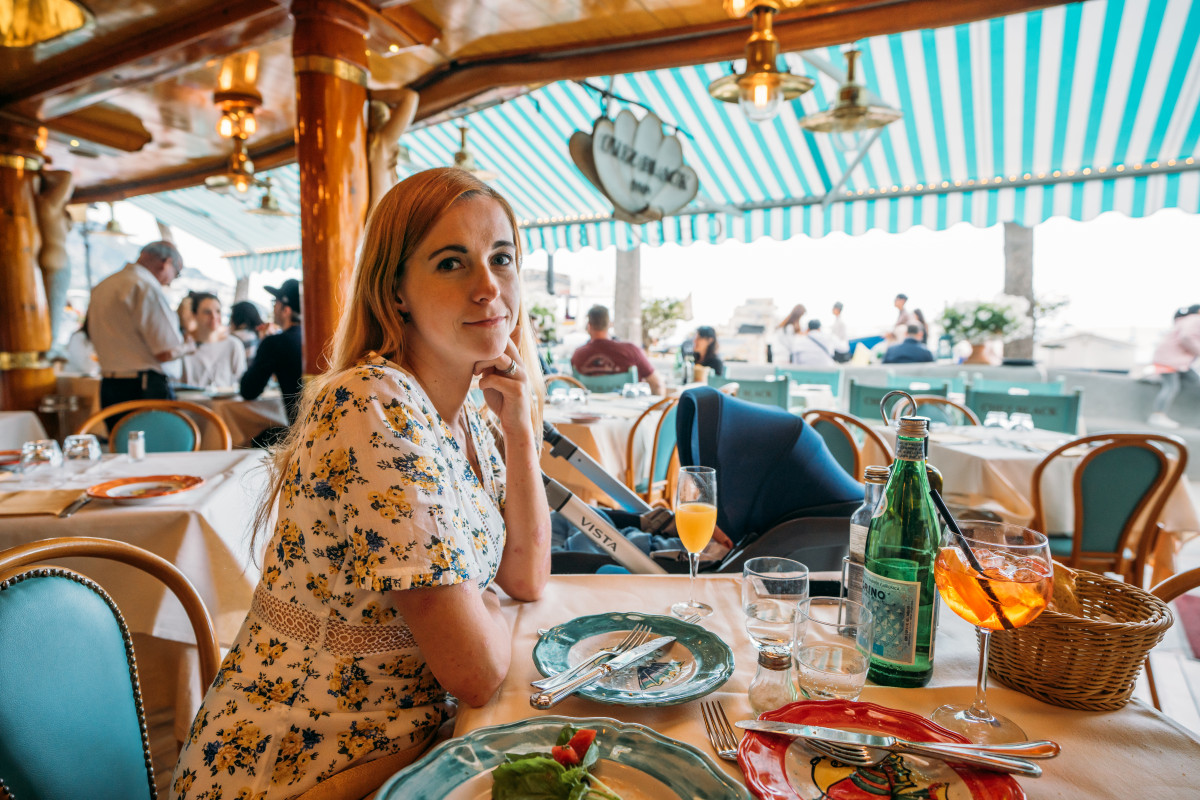 Relaxed dining at an Italian seaside restaurant, with a woman enjoying a meal while traveling with a baby, highlighting family-friendly dining abroad.