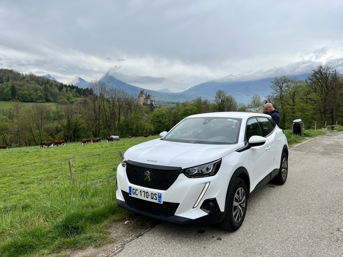 Car parked on a rural road with a backdrop of the French Alps and a castle, illustrating the picturesque drives awaiting when you rent a car in France.
