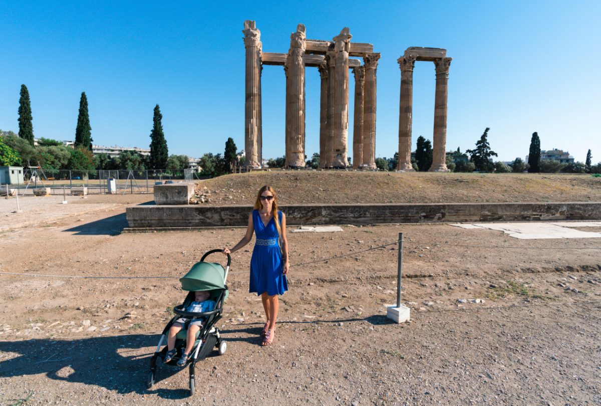 Mother in a vibrant blue dress walks with a stroller at the ancient Greek ruins of the Acropolis, showcasing Athens with a toddler.
