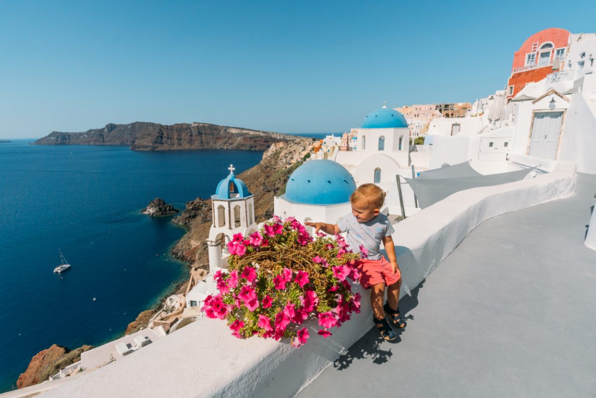 Toddler sitting on a white wall adorned with pink flowers overlooking the iconic Santorini blue domes, a perfect postcard from Greece with a toddler.
