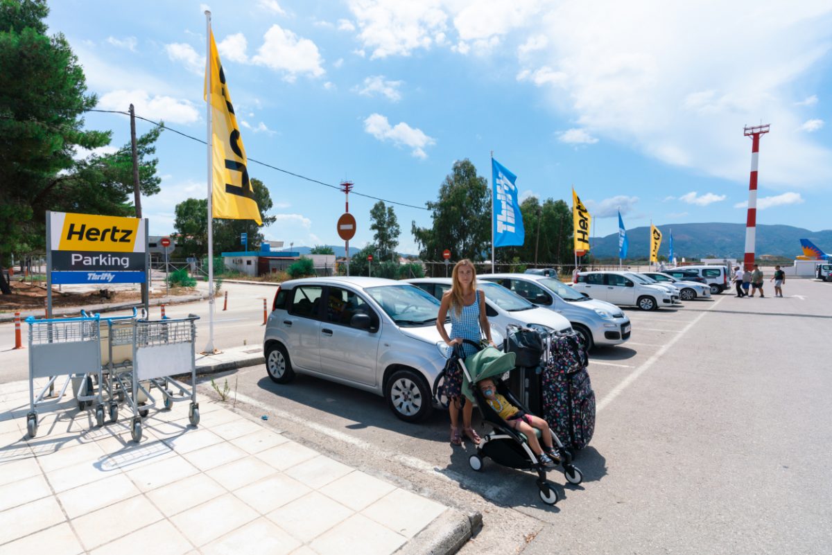 Mother preparing to load a stroller into a car at a Greek rental service, the start of a family adventure with a baby.
