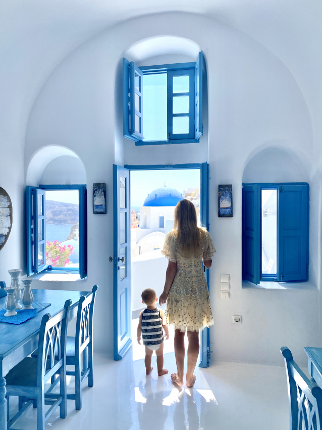 Mother and toddler standing in a white-washed room with a view of the sea, savoring a serene retreat in Greece with a baby.