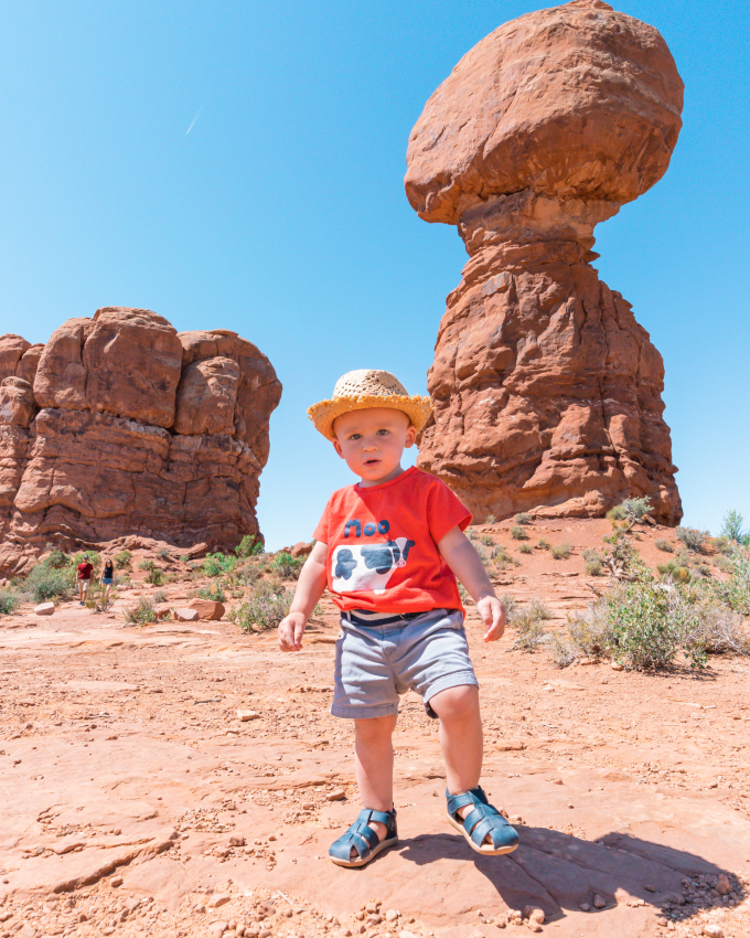 Toddler in a straw hat standing in front of the iconic Balanced Rock at Arches National Park, a must-visit place in the US with a baby