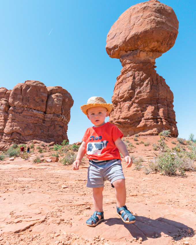 Toddler in a straw hat standing in front of the iconic Balanced Rock at Arches National Park, a must-visit place in the US with baby