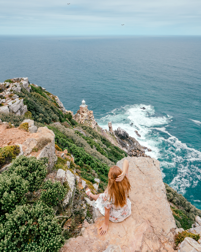 Woman overlooking the historic Cape Point Lighthouse in Cape Town from a rocky cliff, a famous maritime landmark at the tip of the Cape Peninsula.