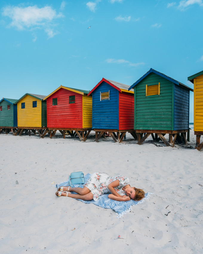 Woman lying on the sand in front of colorful beach huts in Muizenberg, Cape Town, a vibrant and iconic seaside destination for visitors.