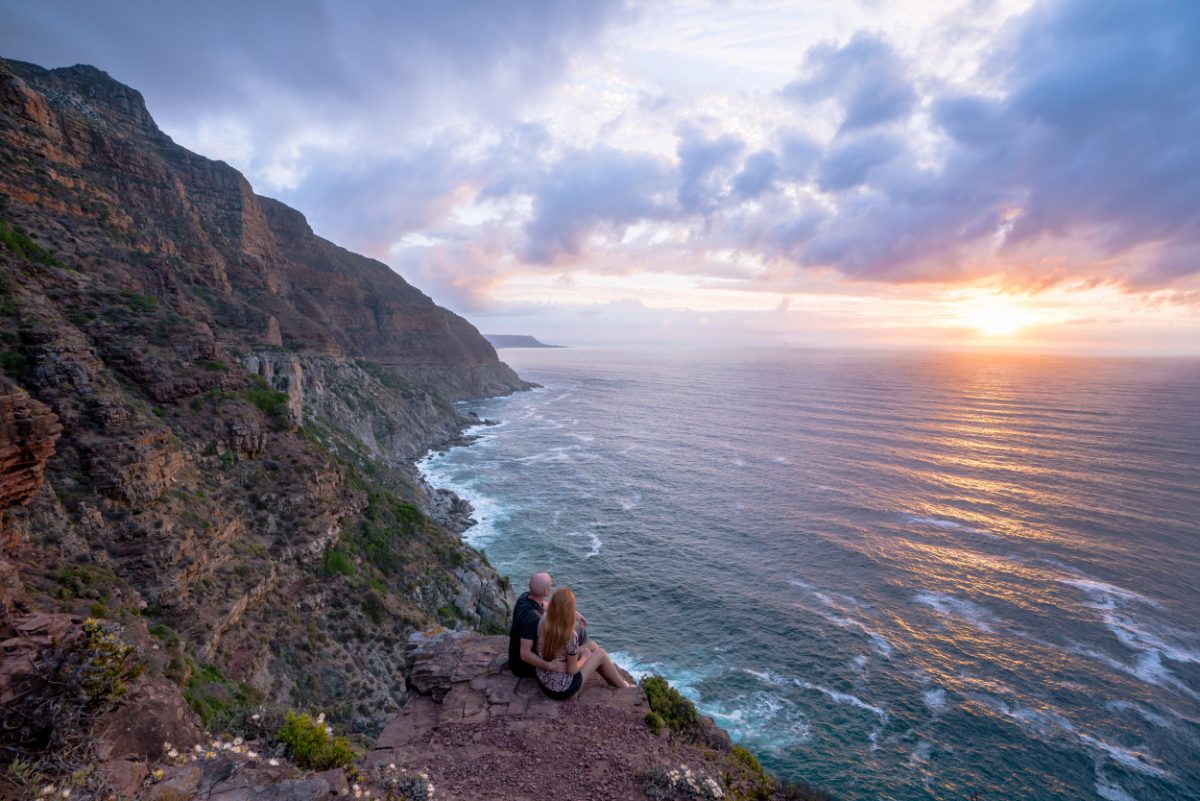 Couple enjoying a romantic sunset view over the ocean from Chapman's Peak Drive in Cape Town, a picturesque spot for end-of-day reflections.