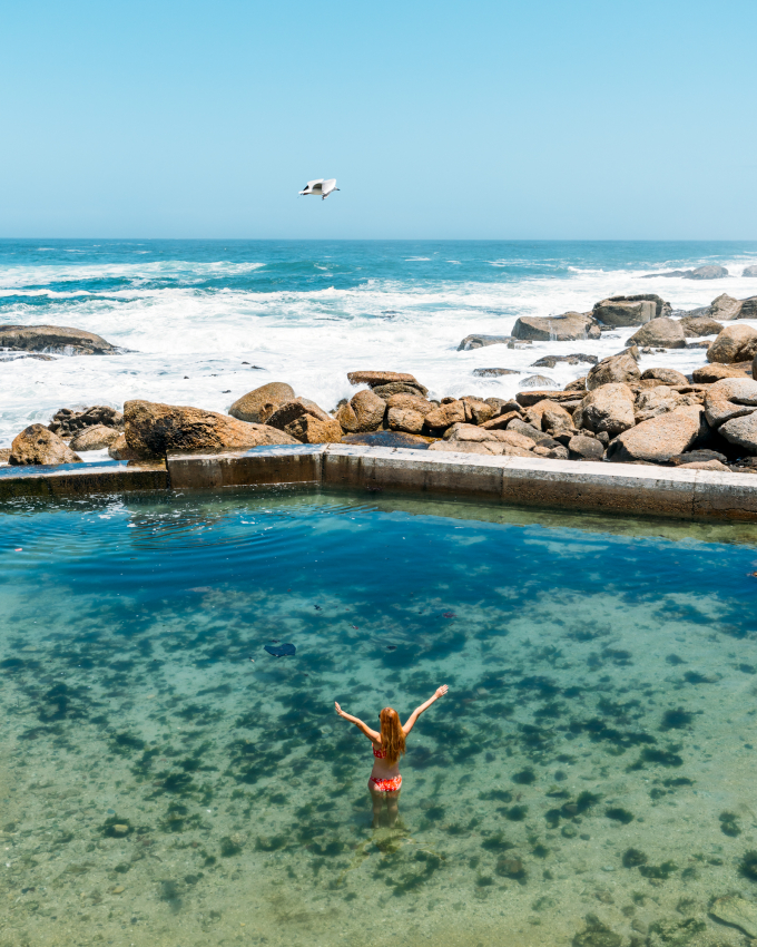 Tourist enjoying the tranquility of a secluded tidal pool in Cape Town, with the waves of the Atlantic Ocean crashing in the background, a hidden gem among the city’s coastal attractions.