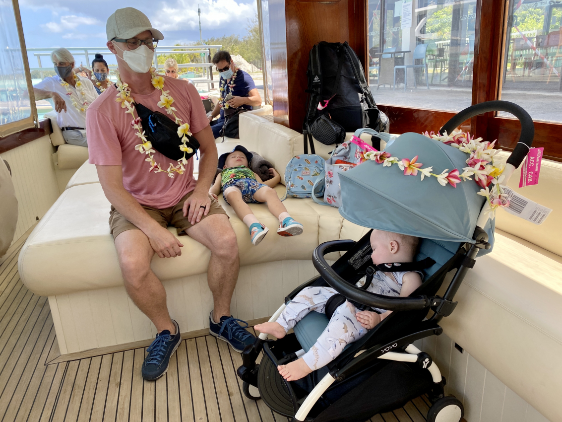 Family on a boat with a sleeping toddler in a portable stroller adorned with a lei, showcasing ease of use during airplane travel.