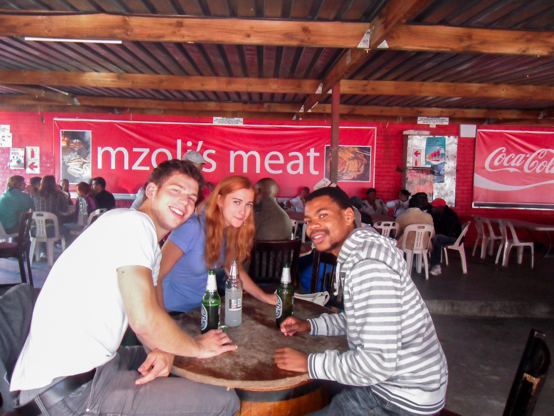 Visitor enjoying a casual dining experience with locals at Mzoli's Meat in Cape Town, offering a taste of everyday life and community spirit in the city.
