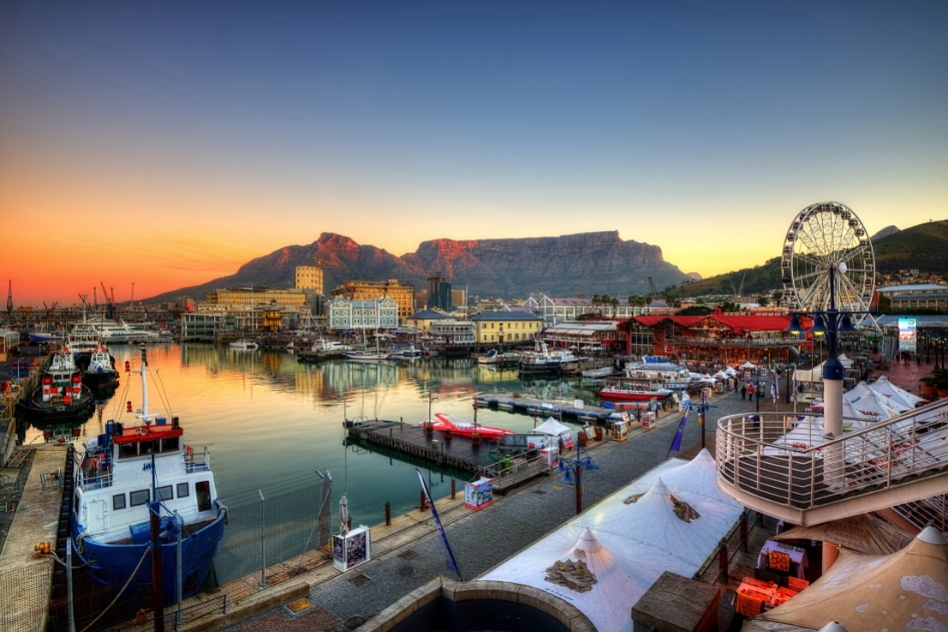 Sunset view of Cape Town's V&A Waterfront with Table Mountain in the distance and reflections on the harbor, a bustling destination for shopping and dining experiences.