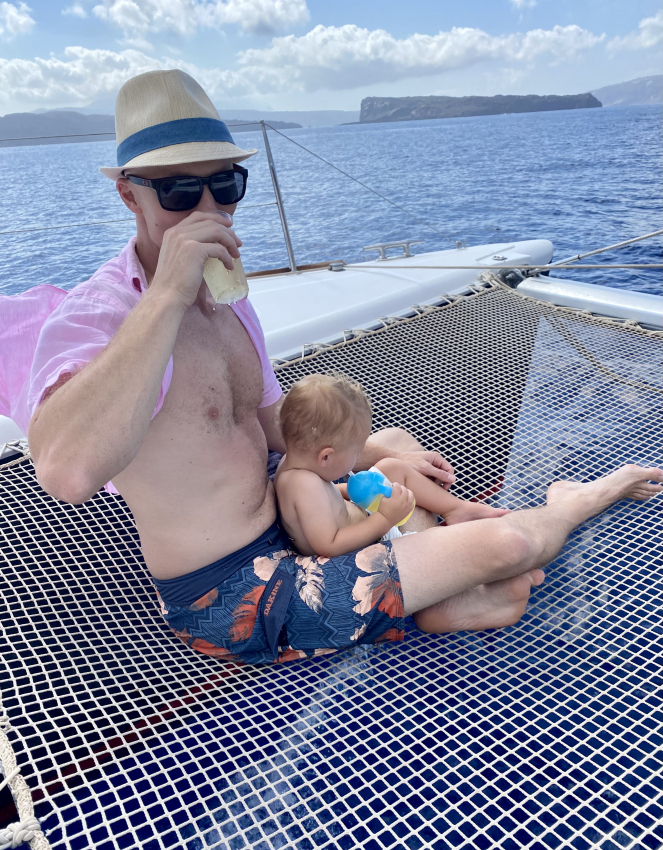 Father sipping a drink while relaxing on a catamaran with his baby in Greece, embodying the best way to travel with a toddler in Greece. 
