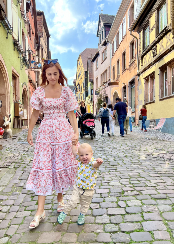 A joyful toddler reaching out happily on a cobblestone street while walking with his mother in a colorful dress, capturing the essence of exploring France with a baby.
