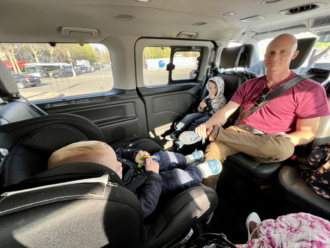 Inside a vehicle, a baby rests in a car seat while a toddler snacks, showcasing the practicalities of road-tripping in France with a baby.
