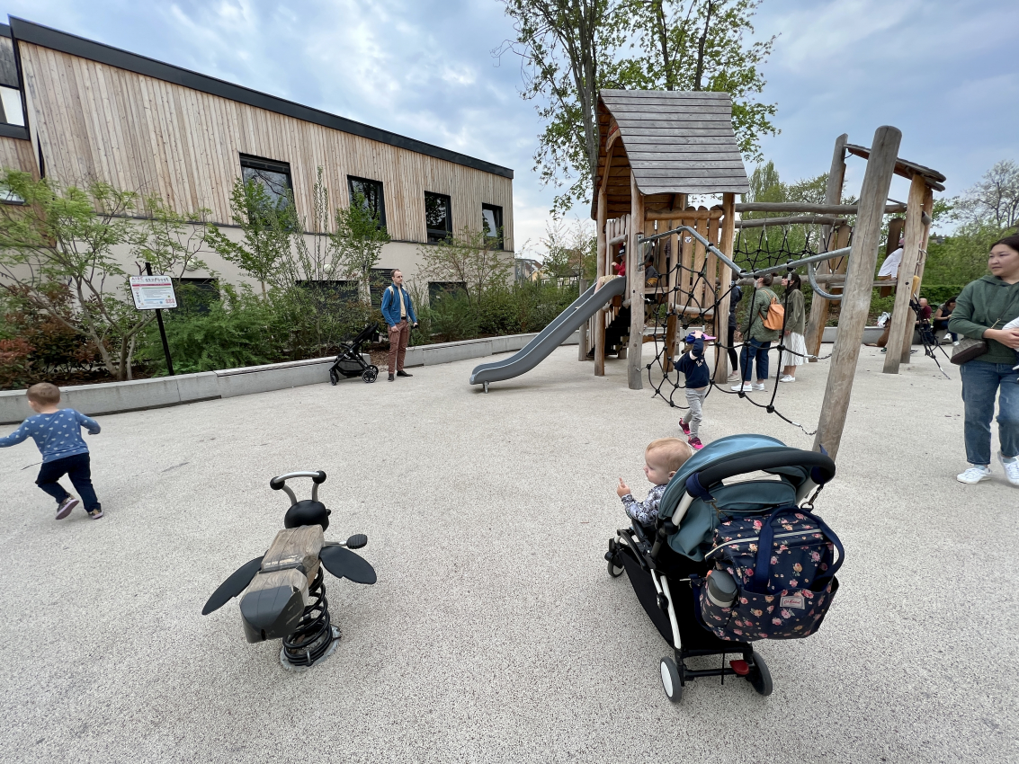 Curious baby peering out from a stroller at a playground in France, depicting the child-friendly side of urban France with a baby.
