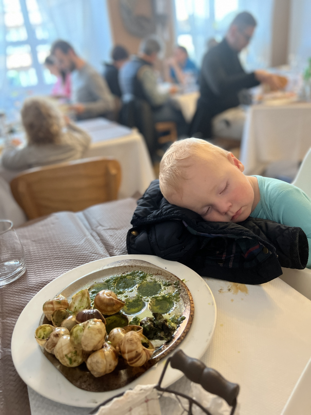 Toddler fast asleep on a restaurant table beside a plate of escargot, illustrating the sometimes exhausting adventures of exploring France with a baby.
