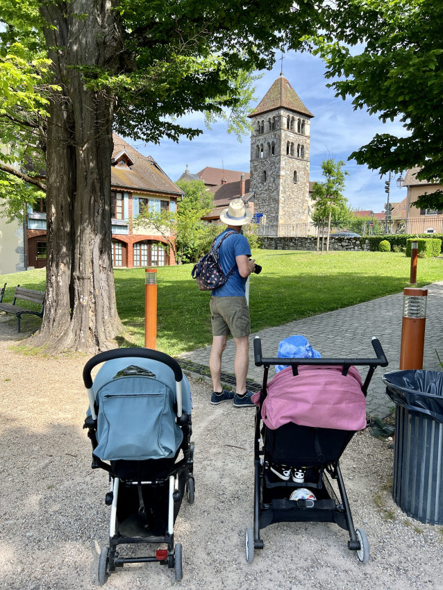 Father and two strollers in a park, with a quaint tower in the backdrop, symbolizing leisurely family walks in France with a baby.
