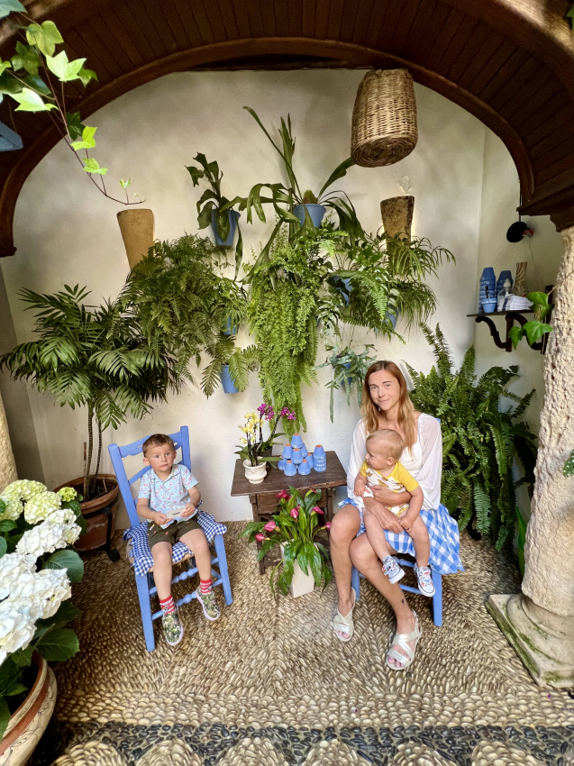 Cozy and lush indoor garden with a mother sitting with her baby and toddler, enjoying the tranquility of a green space in Spain.
