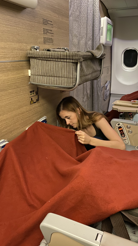 Woman tucked under a red blanket in an airplane seat with a bassinet attached to the bulkhead