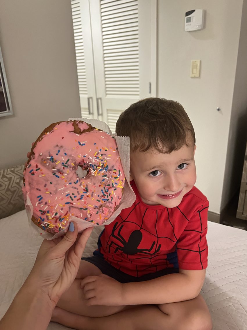 Toddler in a Spider-Man shirt smiling next to an oversized pink donut, enjoying the sweet treats during a family vacation at Universal Studios Orlando with toddlers.
