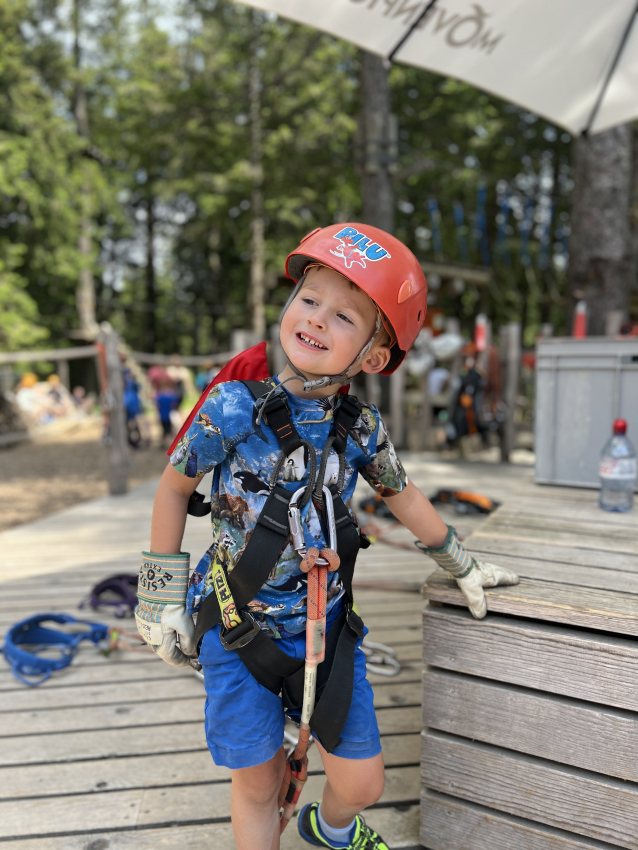 Young boy dressed for adventure, smiling with excitement before a tree-top ropes course, an outdoor family activity in Switzerland.
