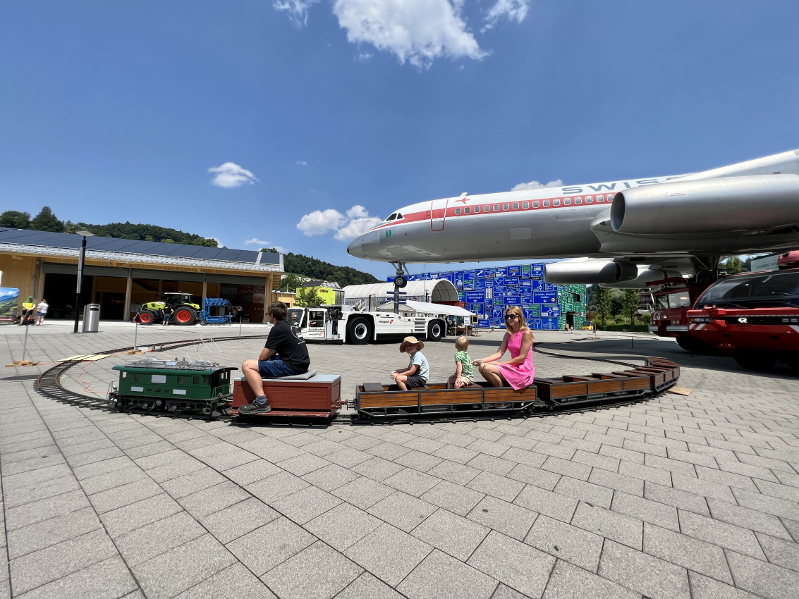 Kids and parents sitting on a miniature train in front of a vintage airplane, a unique experience for a family trip in Switzerland.
